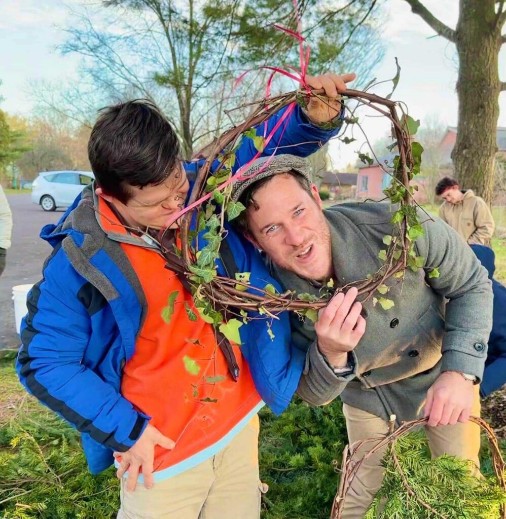 Two people stand in the foreground. One is smiling at the camera and is ducking down to frame their head inside of a wreath. The wreath is being held by the other person, who is smiling down at them and is holding the wreath up above their head.