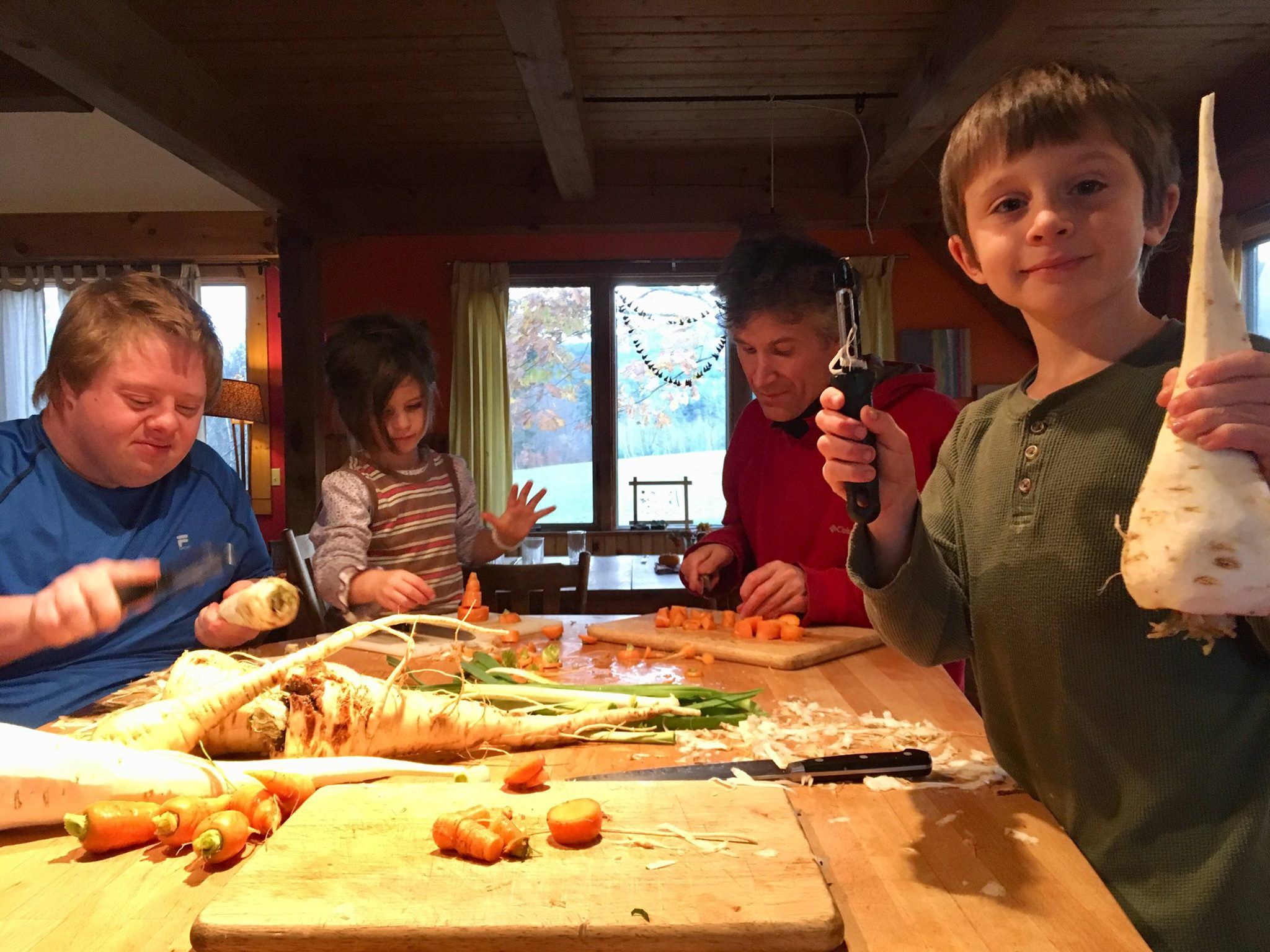 People of all ages peeling and cutting up carrots and parsnips. A child in the foreground holds up a large parsnip with pride, while another in the background is stacking up carrot slices into a tower.