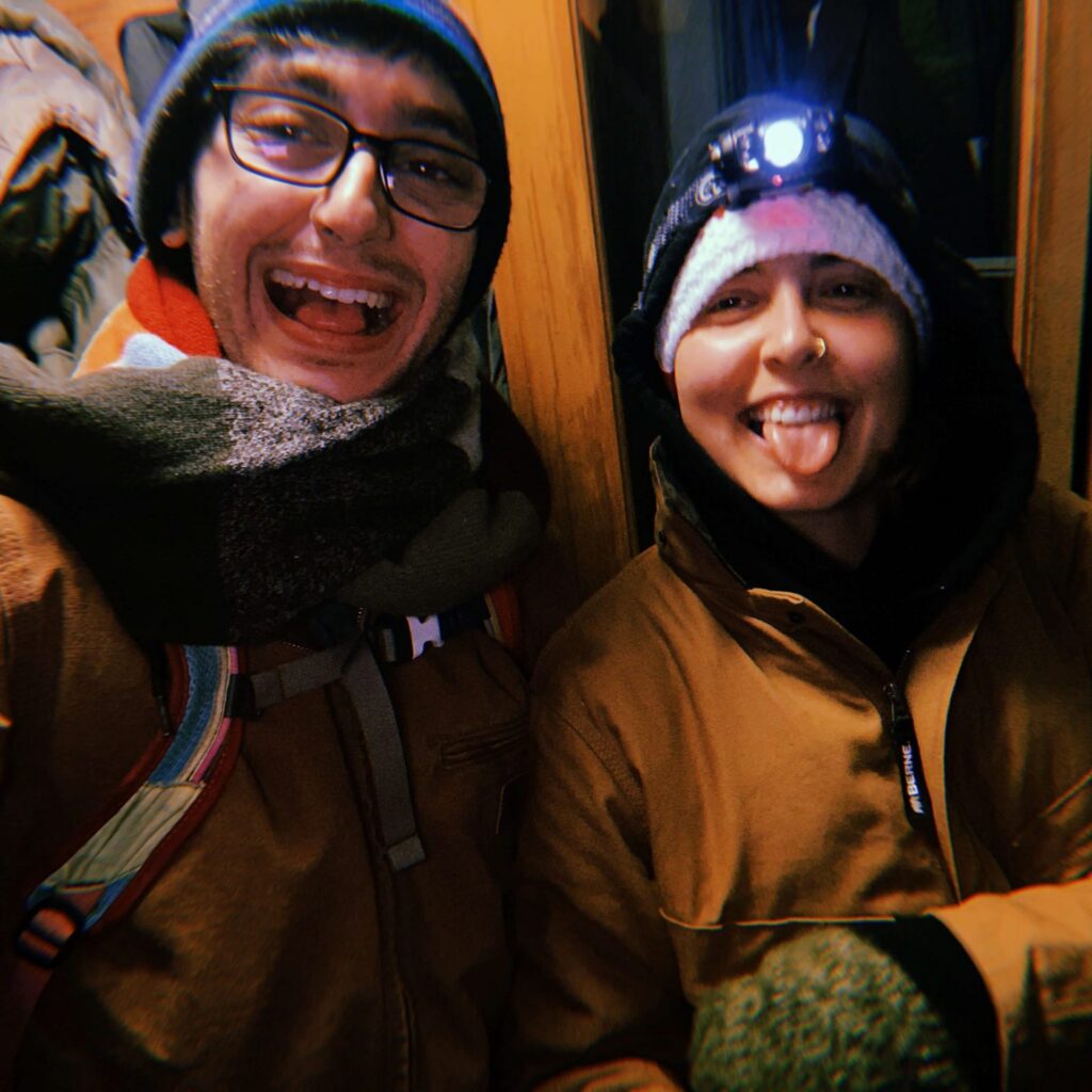 Two people in winter clothing smiling for the camera. One wears a lit headlamp.