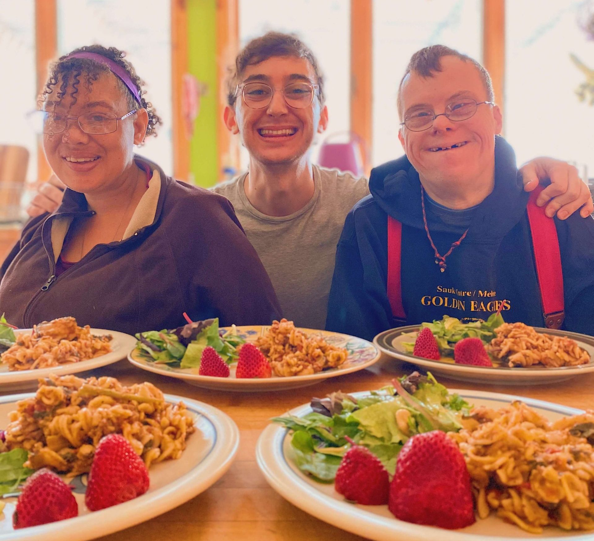 Nicolo sits between two friends at a table, all three are smiling, and Nicolo's arms are around the friends. In front of them are five plates of food.
