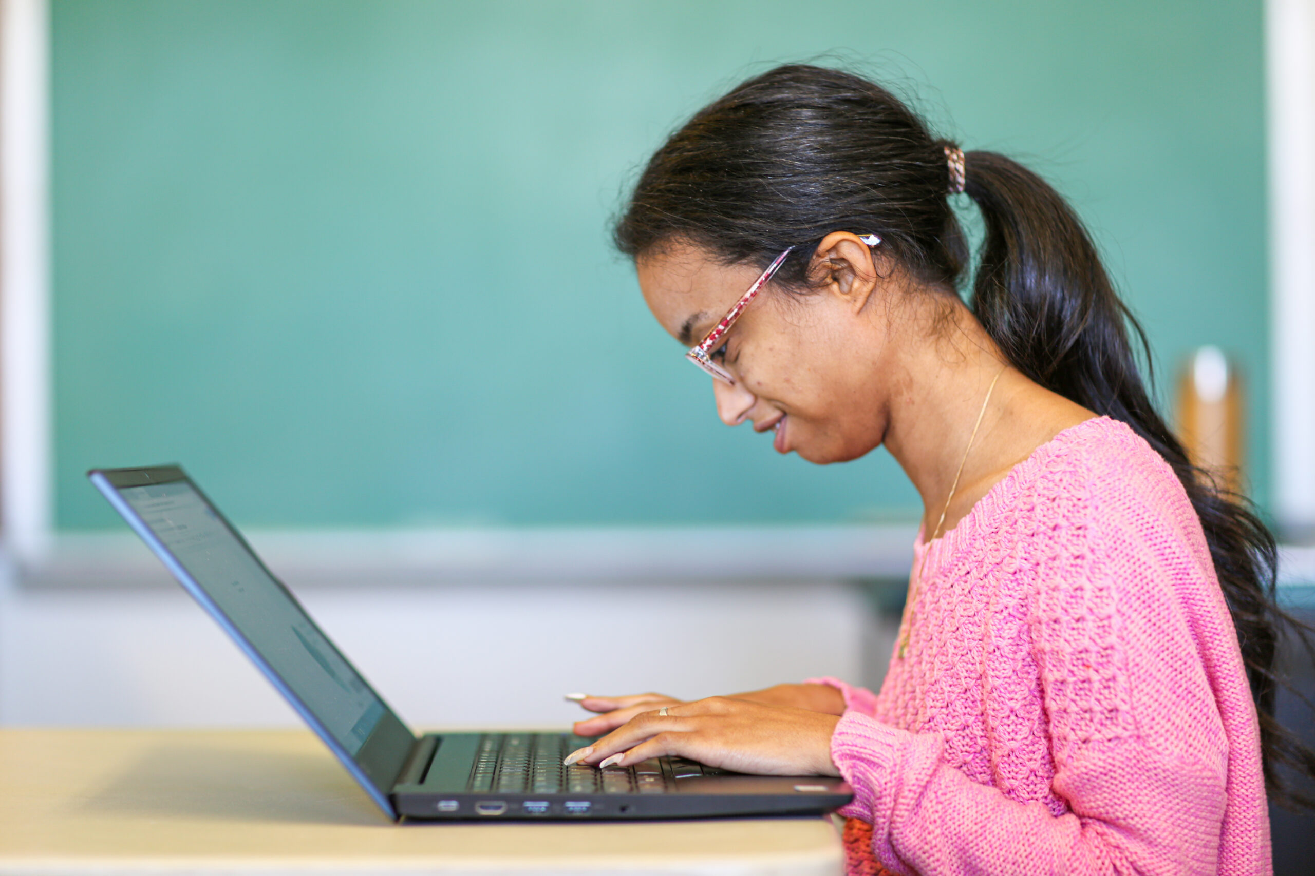 A young woman in a pink sweater types on a laptop in a classroom