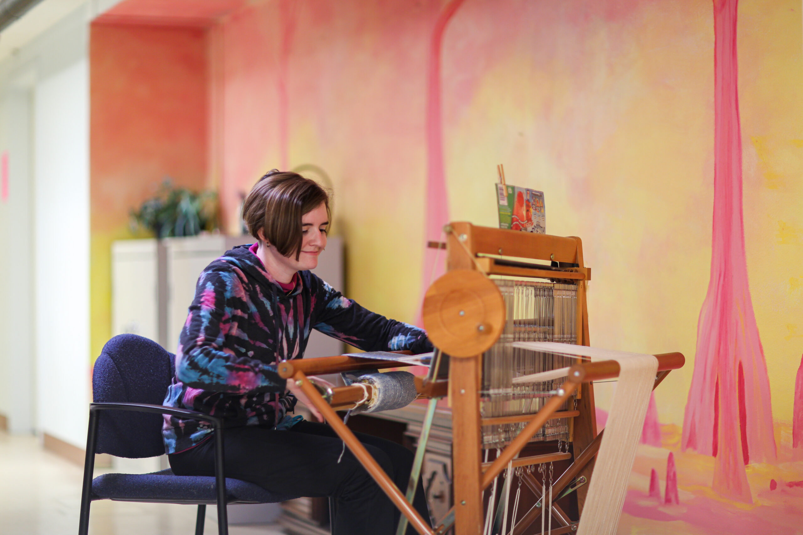 A young adult in a tie-dye hoodie weaves on a small floor loom