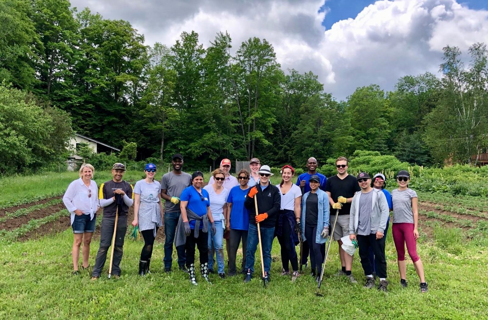 A group of adults standing together and smiling in front of a garden, holding their gardening tools and wearing hats and sunglasses.
