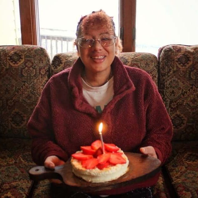 Sara is wearing a red sweatshirt over a white tshirt. She is smiling while holding a cake with white icing, strawberries, and a single lit candle.