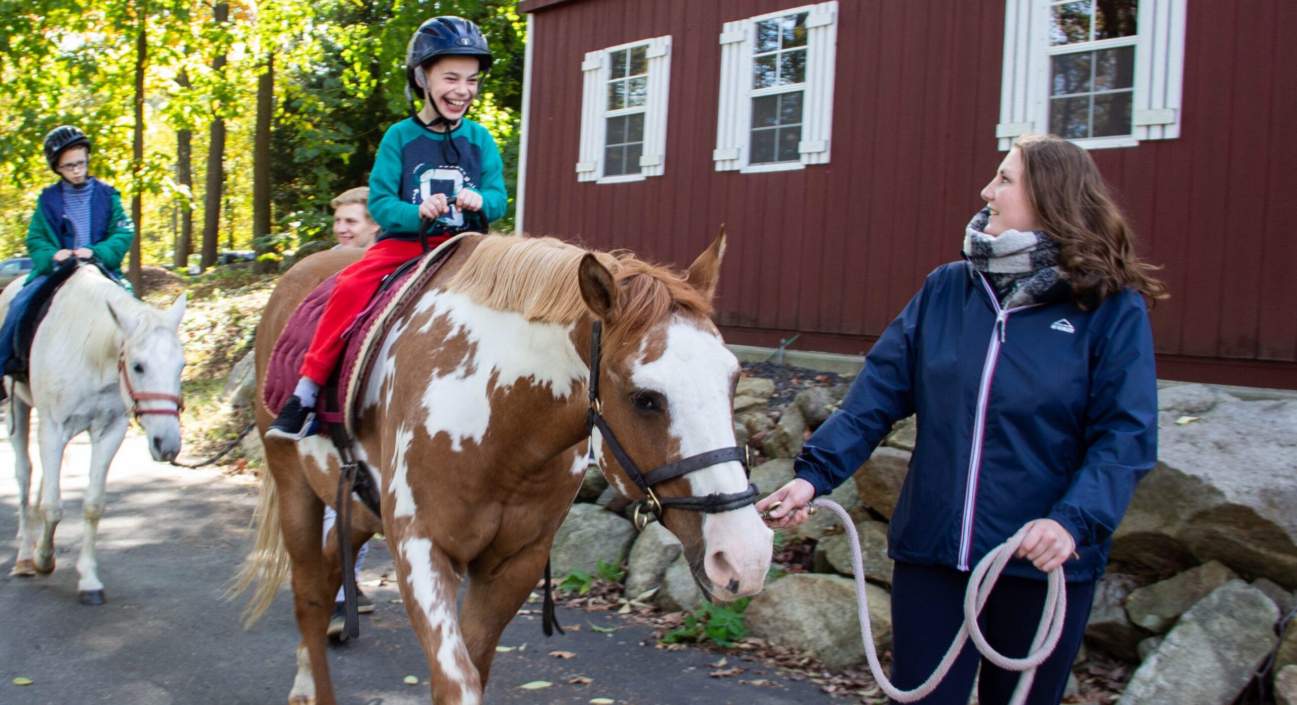 Two children in helmets riding horses guided by adults. The boy in front is laughing with delight.