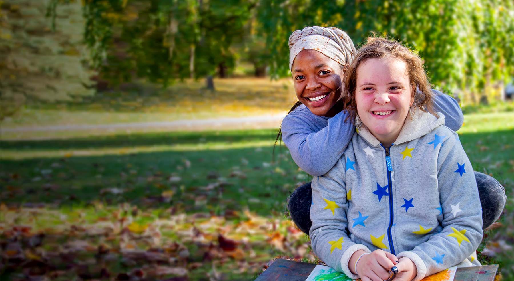 Two women smile at the camera, sitting outdoors in the sunshine.
