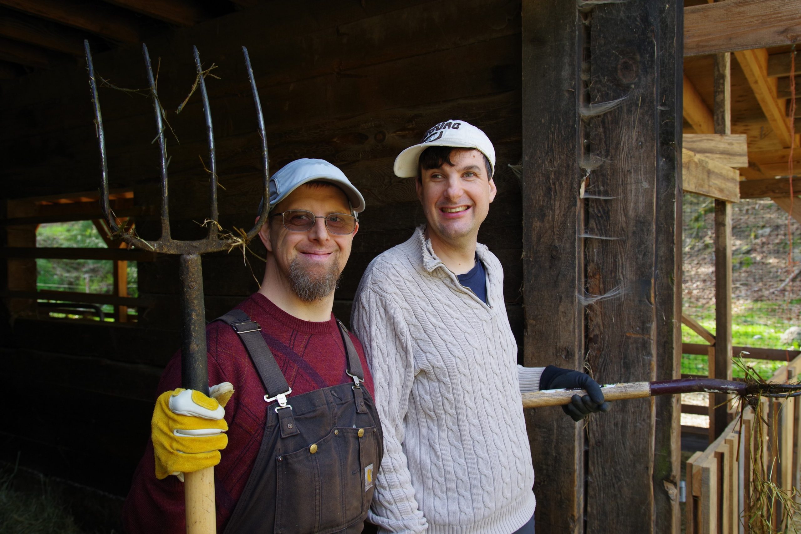 Two men standing in a barn, one holding a pitchfork