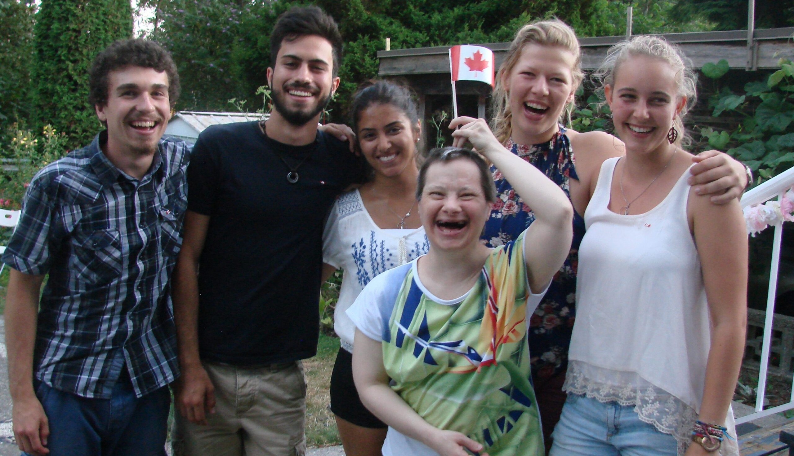 A group of people laughing together and hugging. One person holds up a Canadian flag with delight.