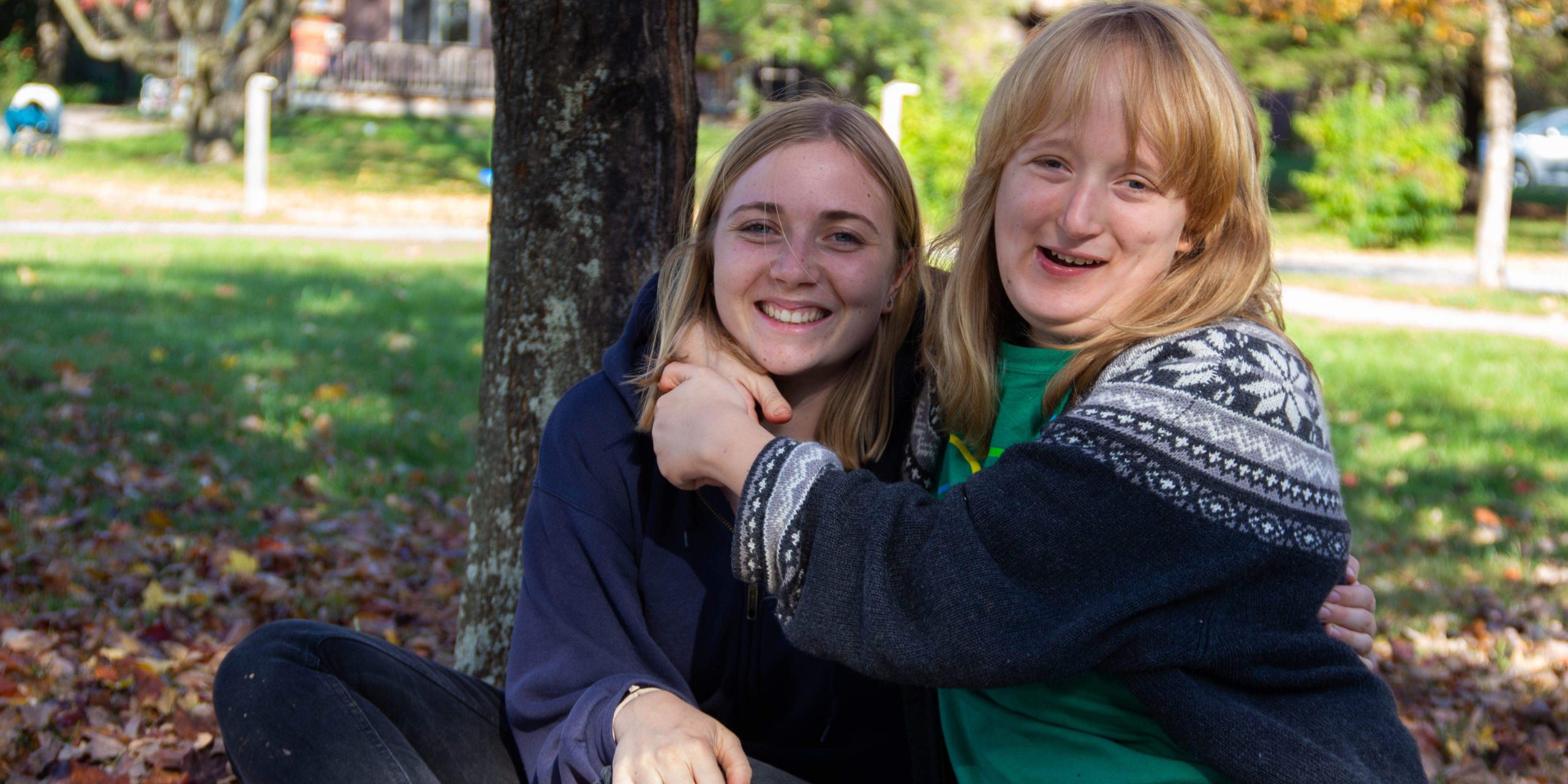 Two girls hugging each other outside under a tree and smiling at the camera