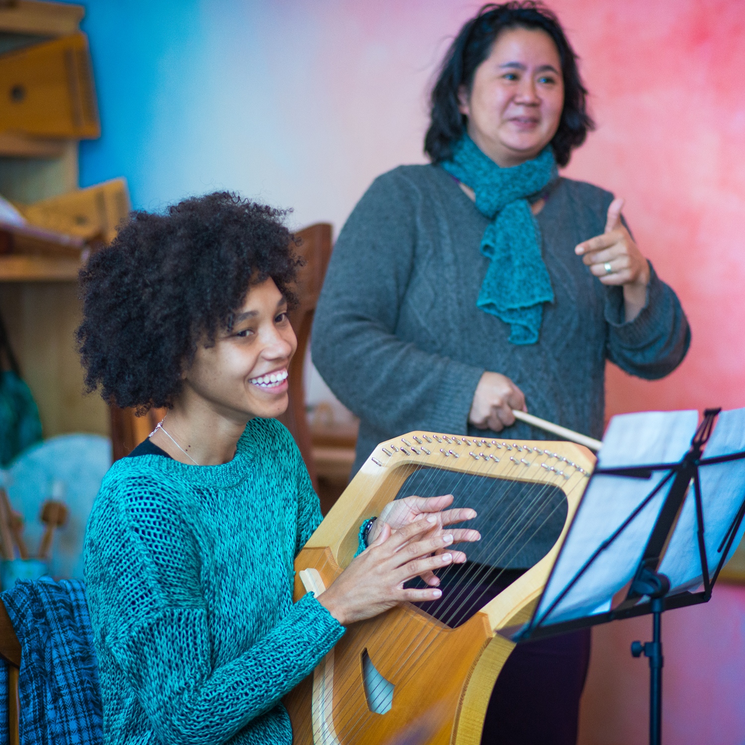 A woman plays a lyre as another woman conducts in the background