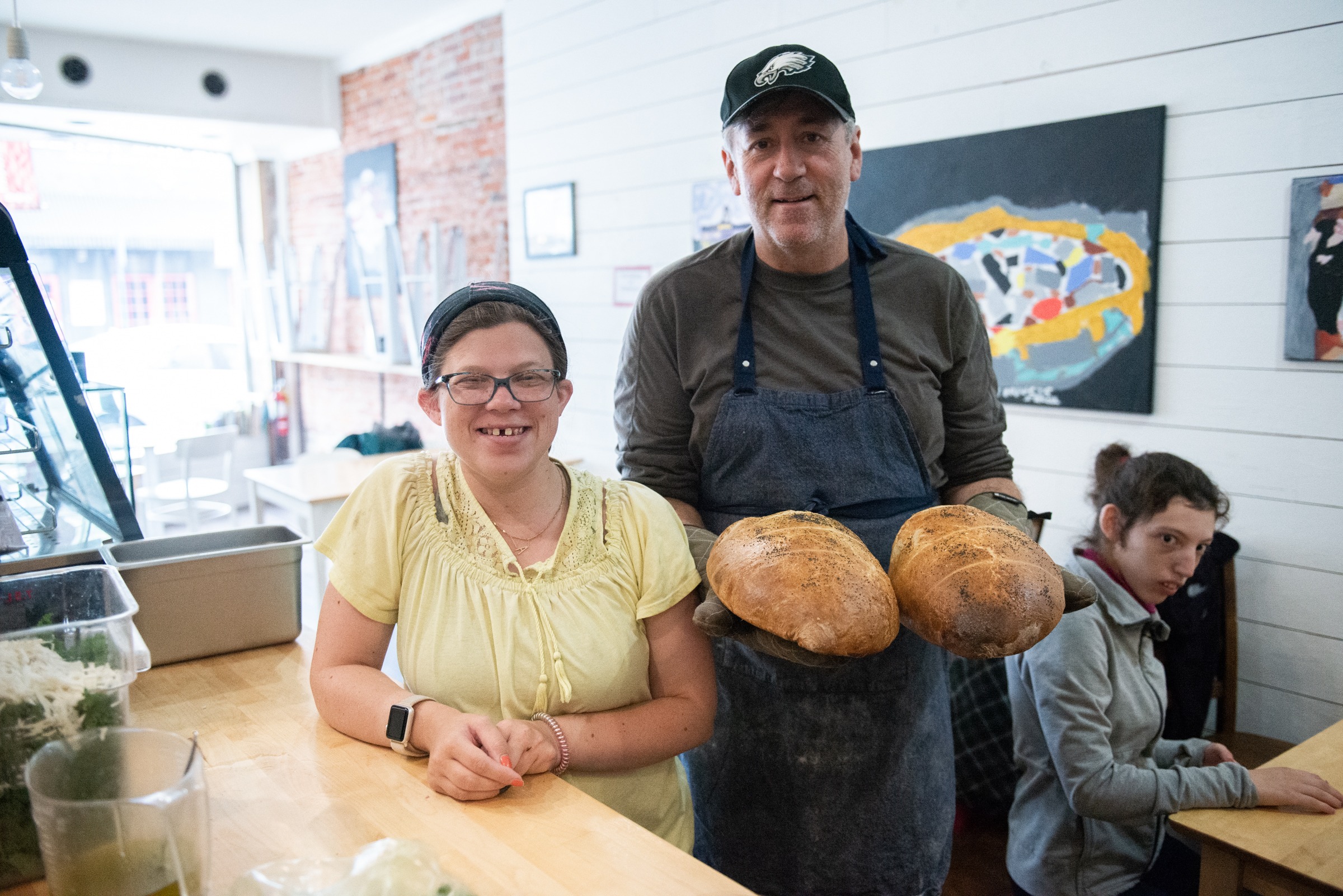 Three people in a bakery, one of whom is holding up two freshly-baked loaves of bread.