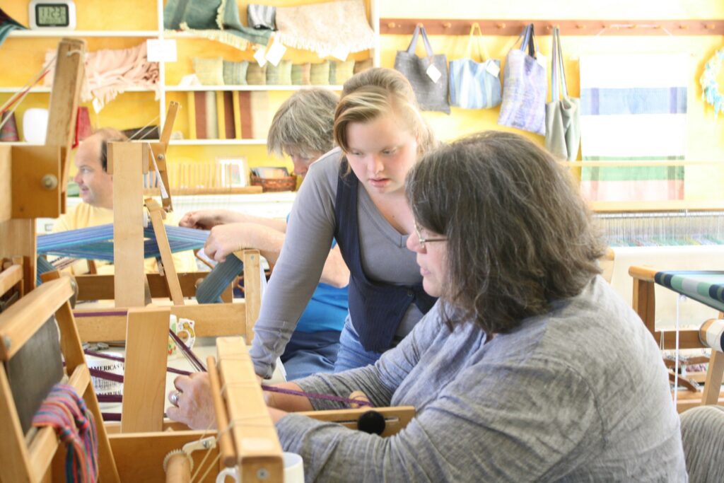 Three people working together: one weaving on a floor loom, one weaving on an inkle loom, and one assisting.