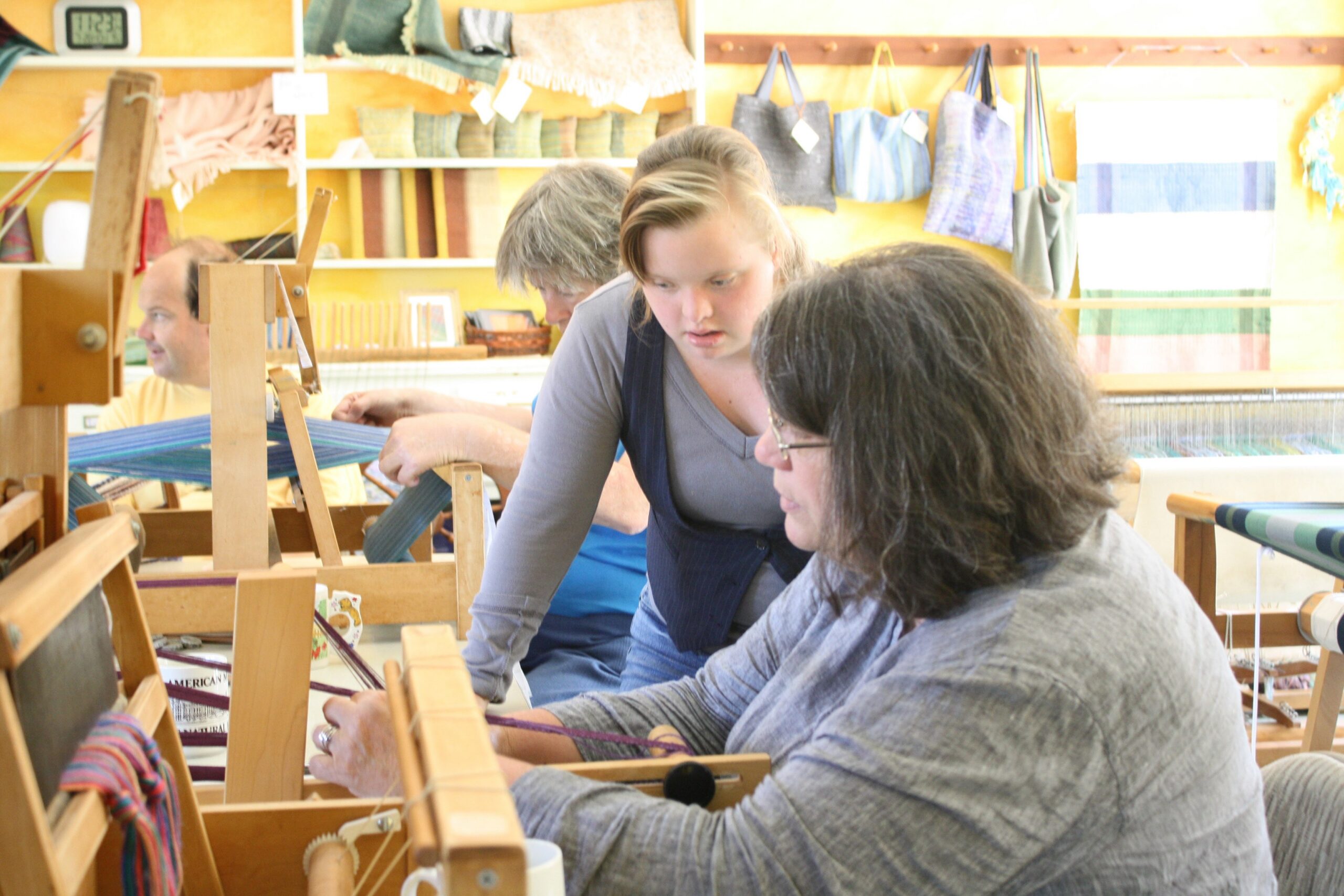 Three people working together: one weaving on a floor loom, one weaving on an inkle loom, and one assisting.