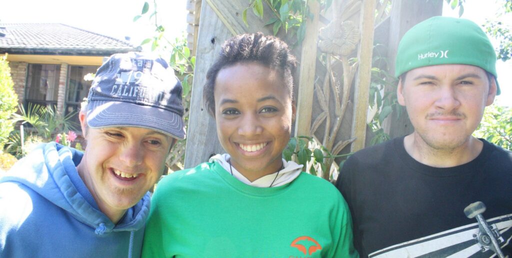 Brittany smiles while standing between two friends at Camphill. They all have their arms around each other. On the left is a friend wearing a blue hoodie and a blue baseball cap. On her right is a friend wearing a black shirt with a skateboard on it and a green baseball cap worn backwards. Brittany is wearing a green camphill t-shirt over a white hoodie.