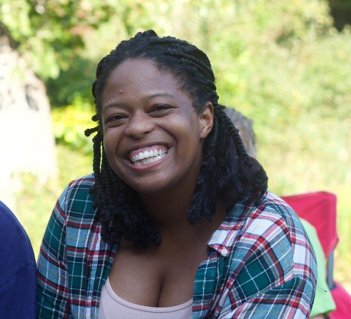 Gabi is smiling for the camera. She is wearing a pink tank top and a green and pink flannel. She is outside, and there is greenery in the background.