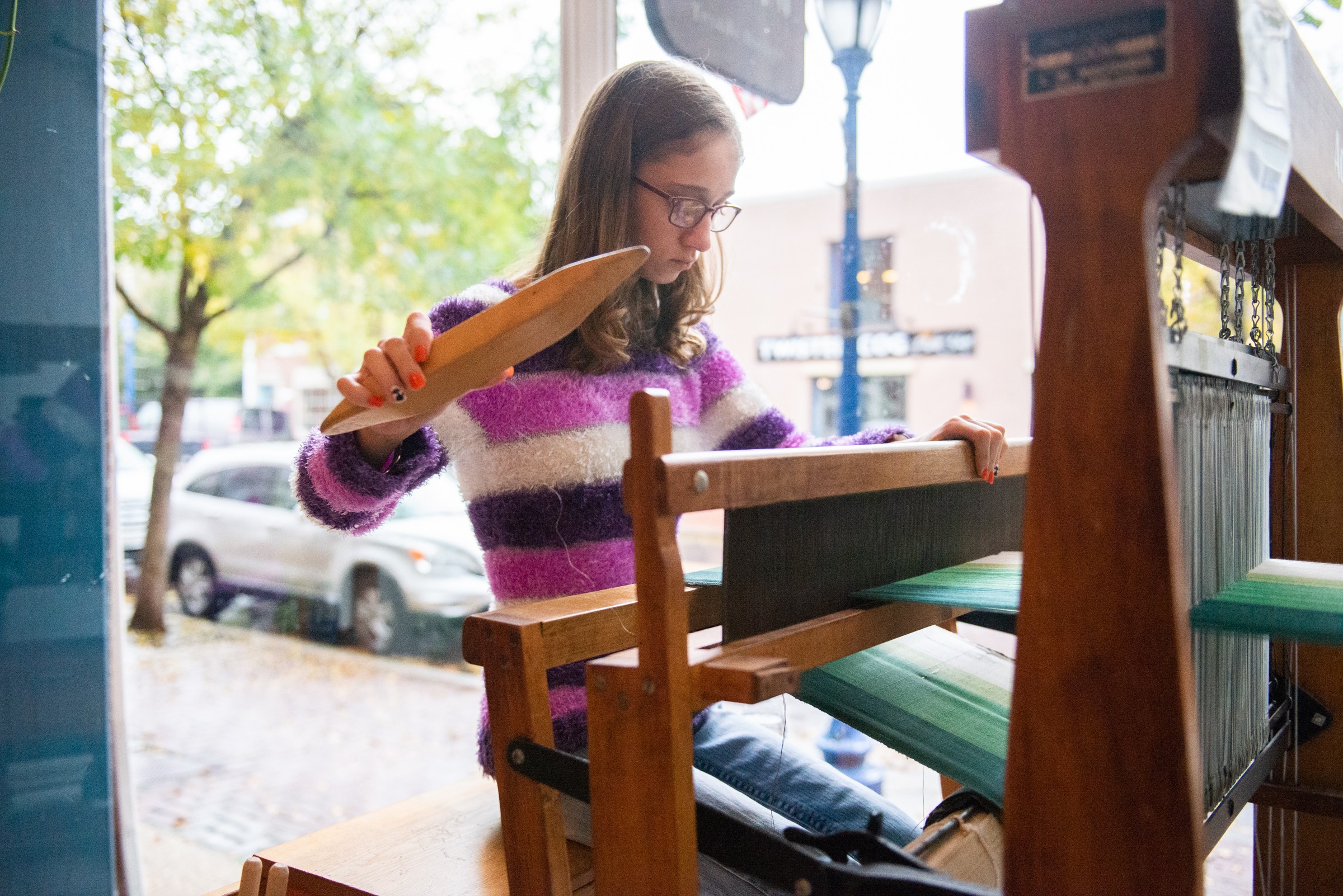 A young woman in a striped purple sweater, weaving a green gradient fabric on a floor loom.