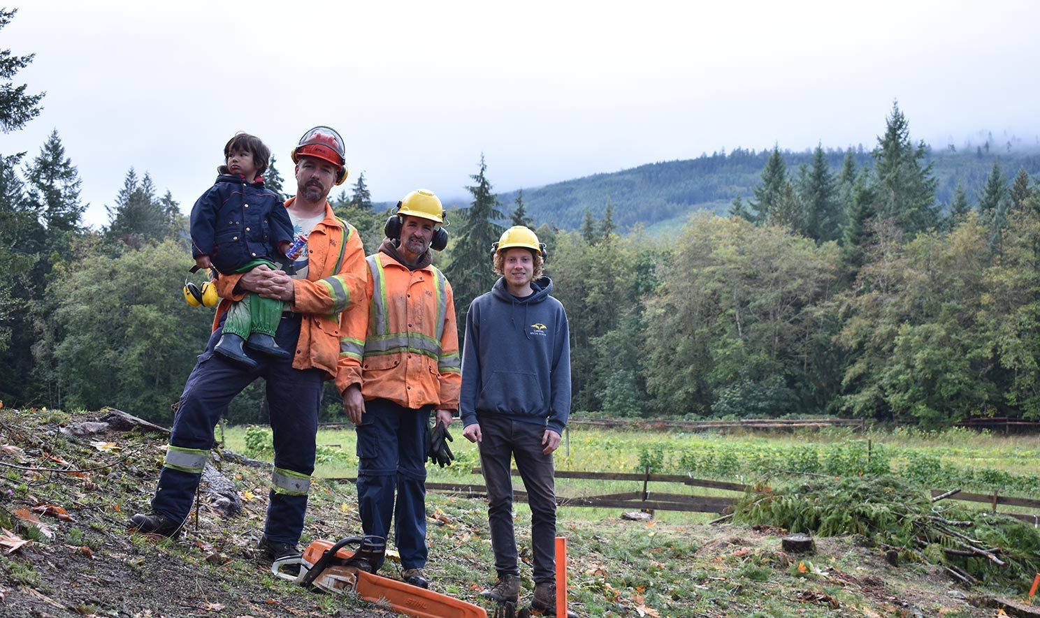 Three men and a toddler stand posing for the camera over their forestry tools.