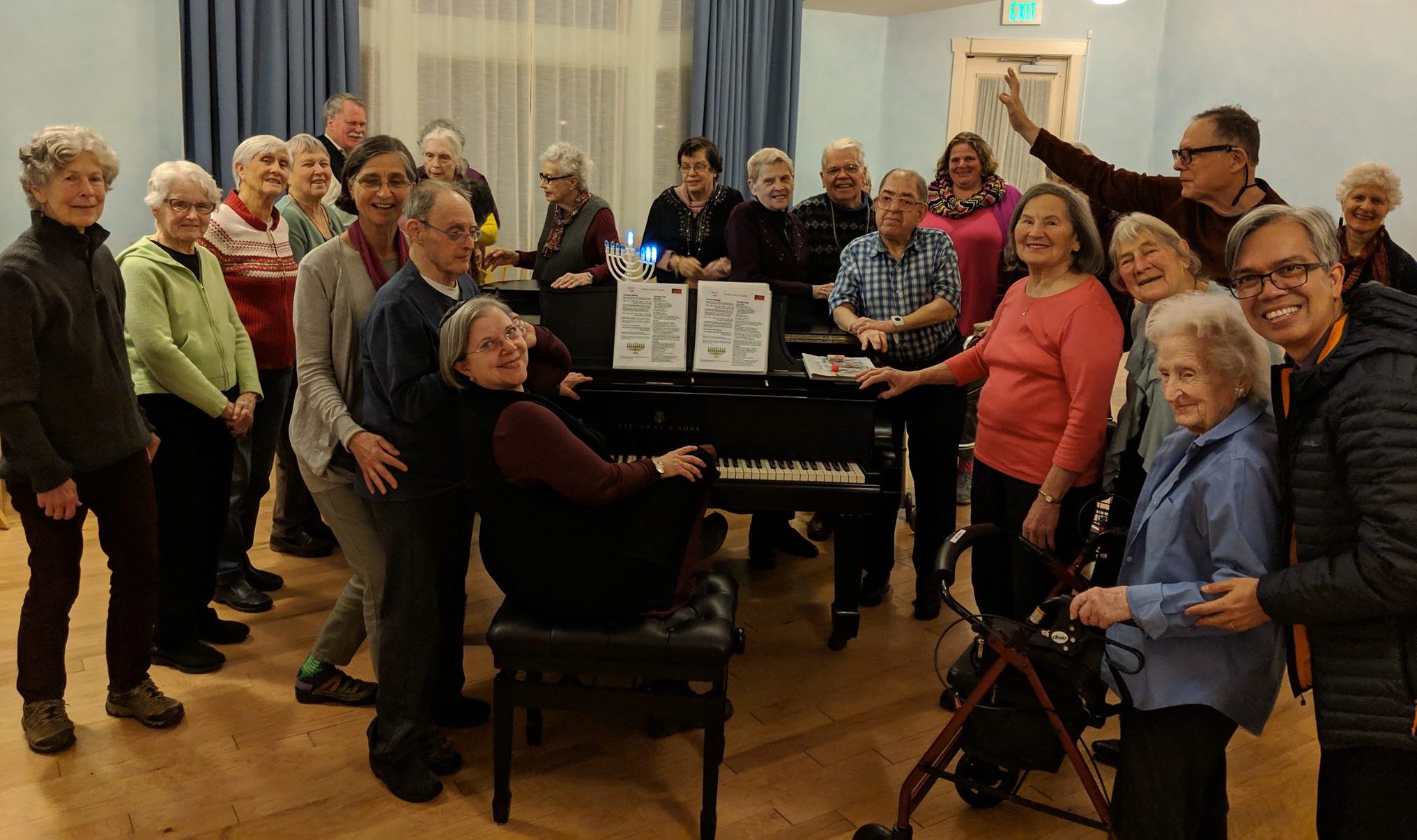 Older adults surround a piano with an electric menorah on it, and the pianist turns backwards to smile at the camera with them.