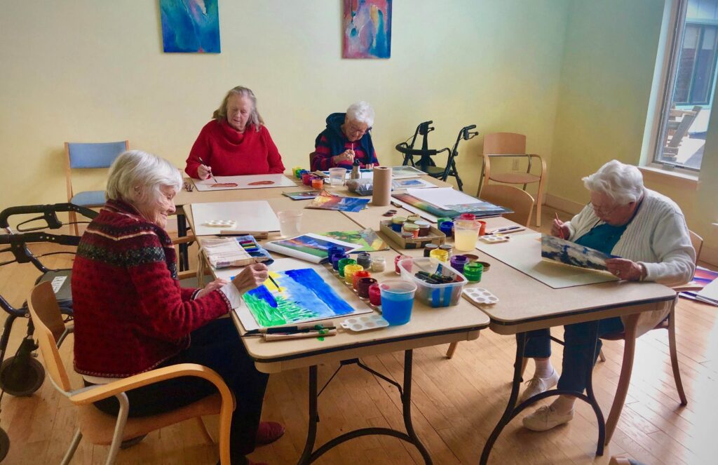 Elderly women sit around two craft tables, painting nature scenes.