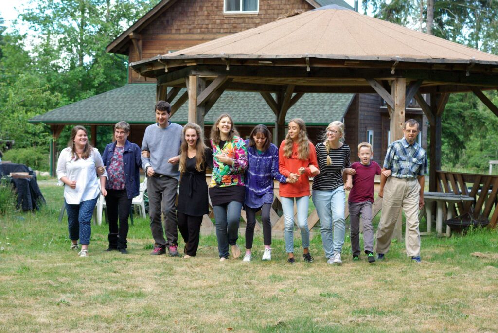 A group of people of all ages walk together in a line outdoors by an old brown house.