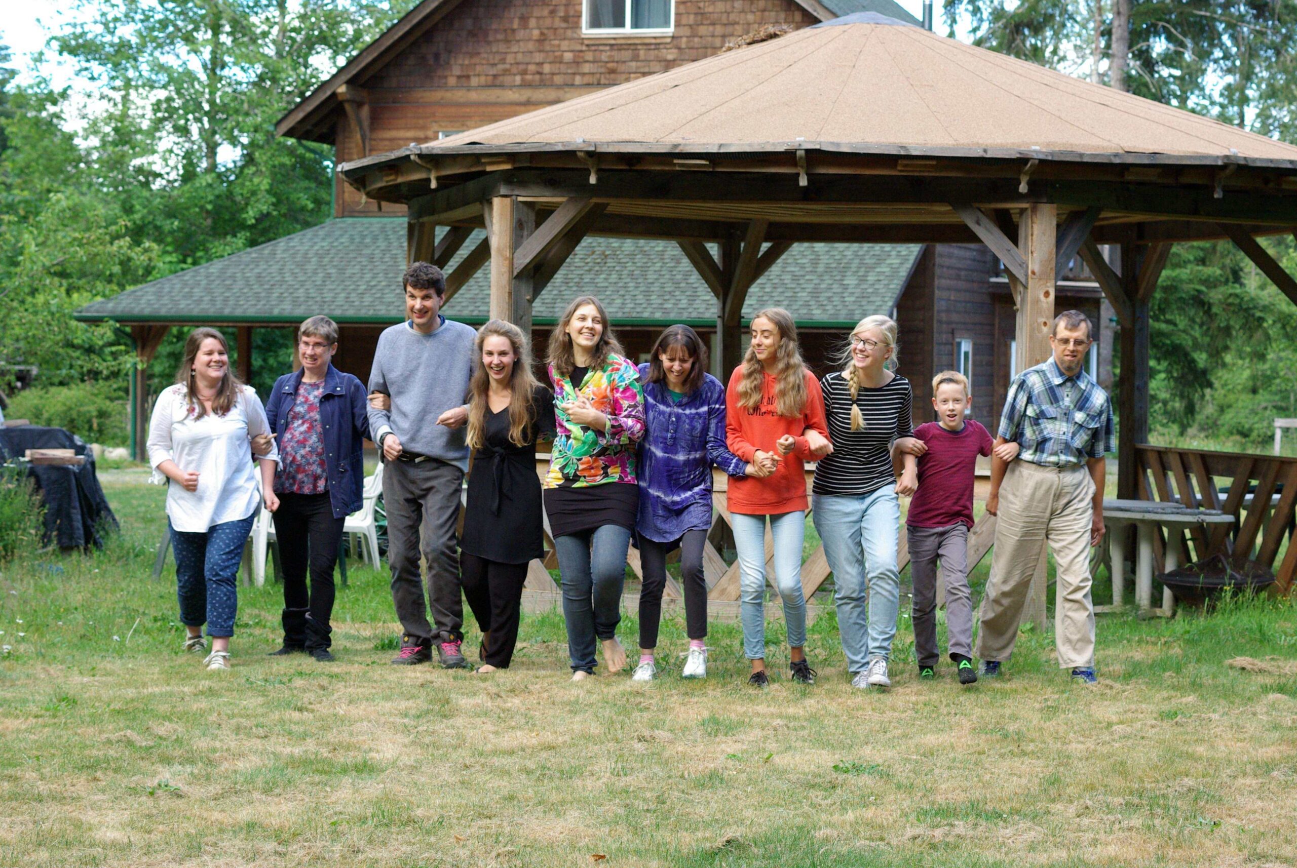 A group of people of all ages walk together in a line outdoors by an old brown house.