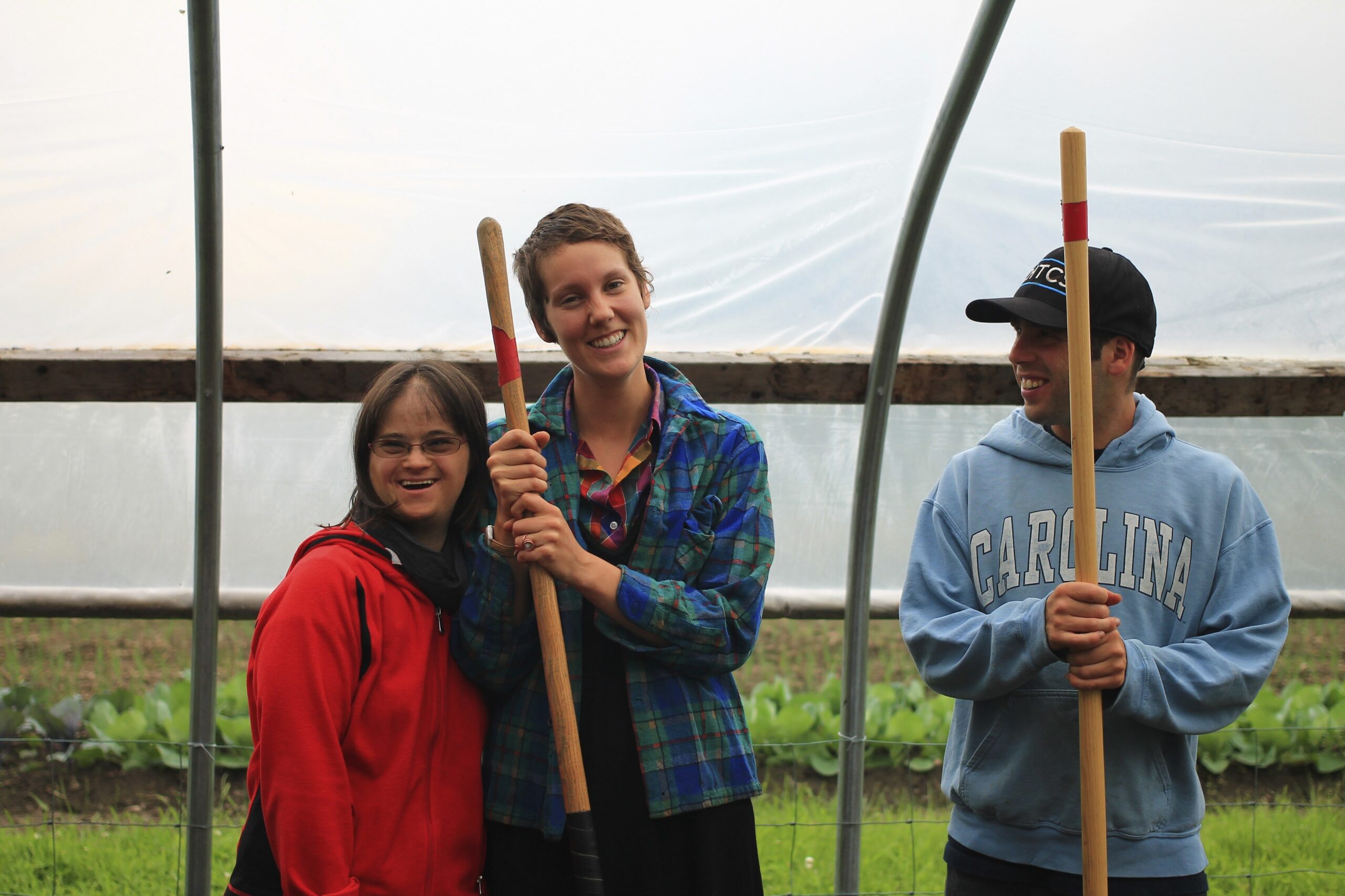 Three people standing together in a greenhouse and smiling, two of them holding gardening tools.