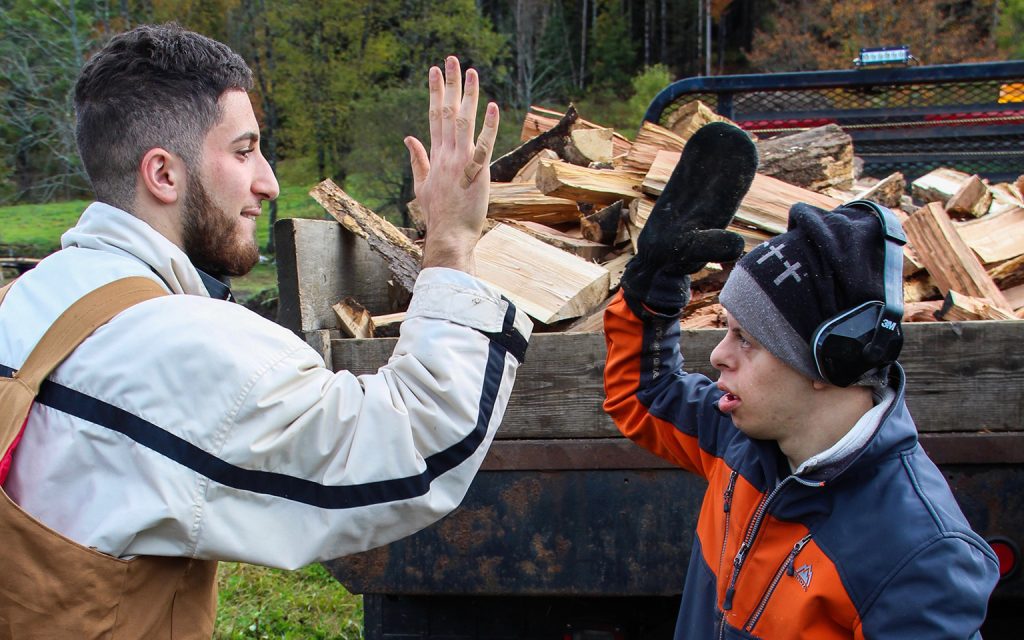 Two young men high-five in front of a truck filled with chopped wood.