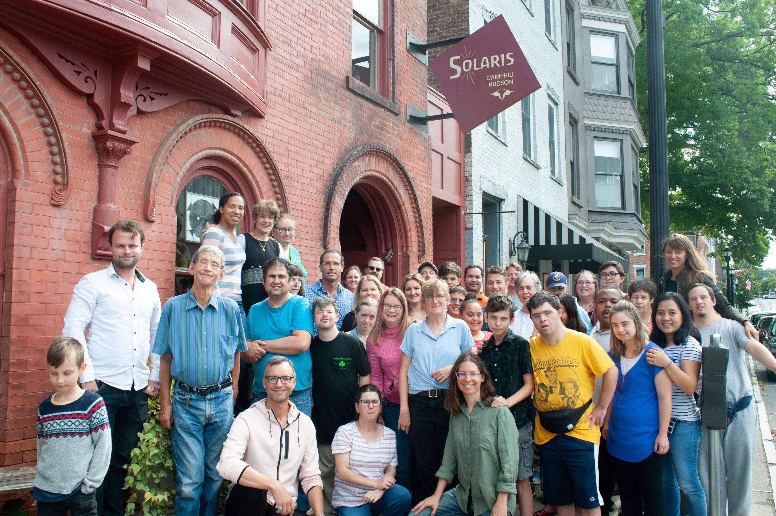 A group of people of all ages pose together under the sign for Solaris Camphill Hudson