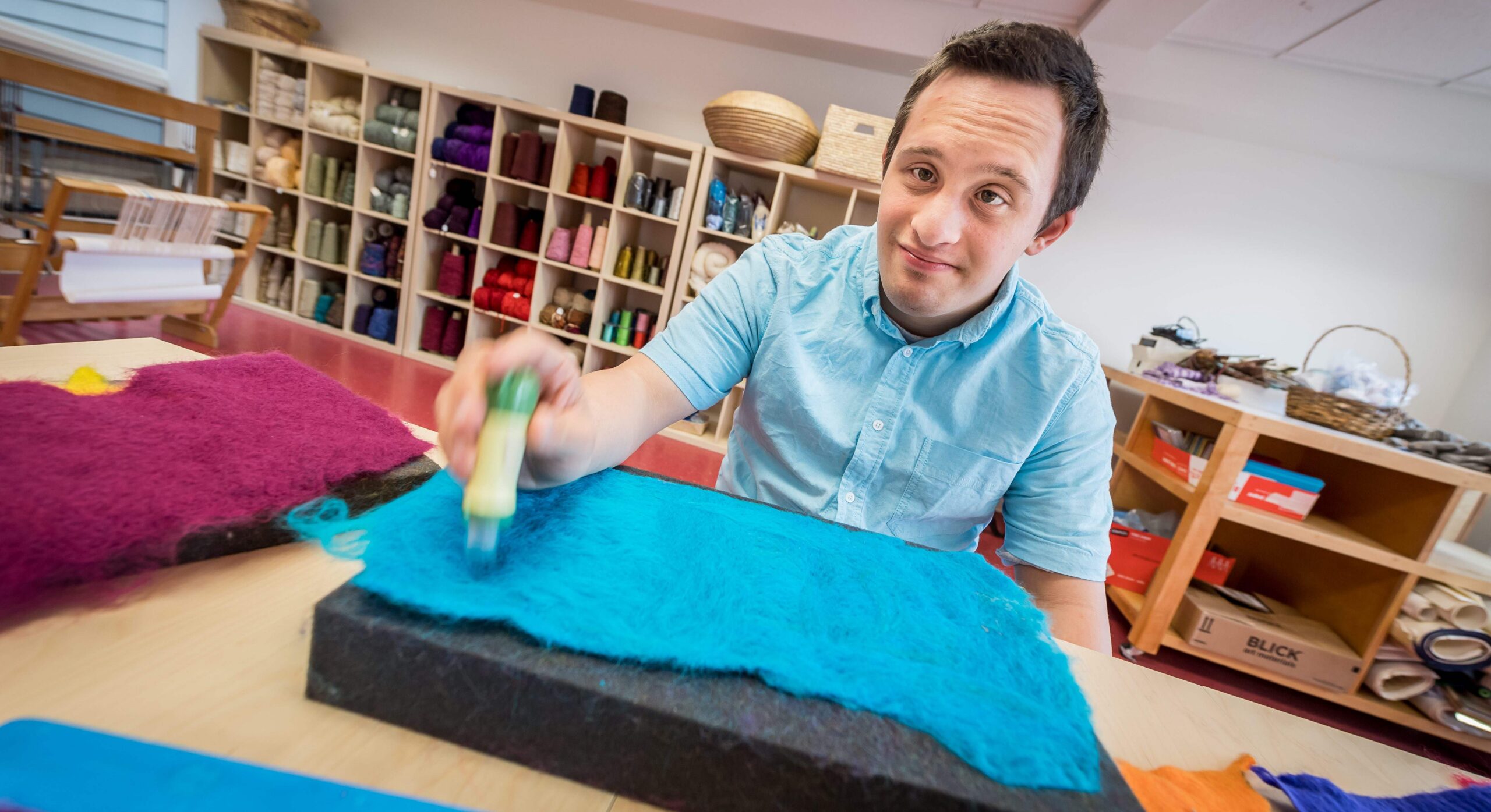 A young man felting blue fibers in a workshop filled with craft materials.