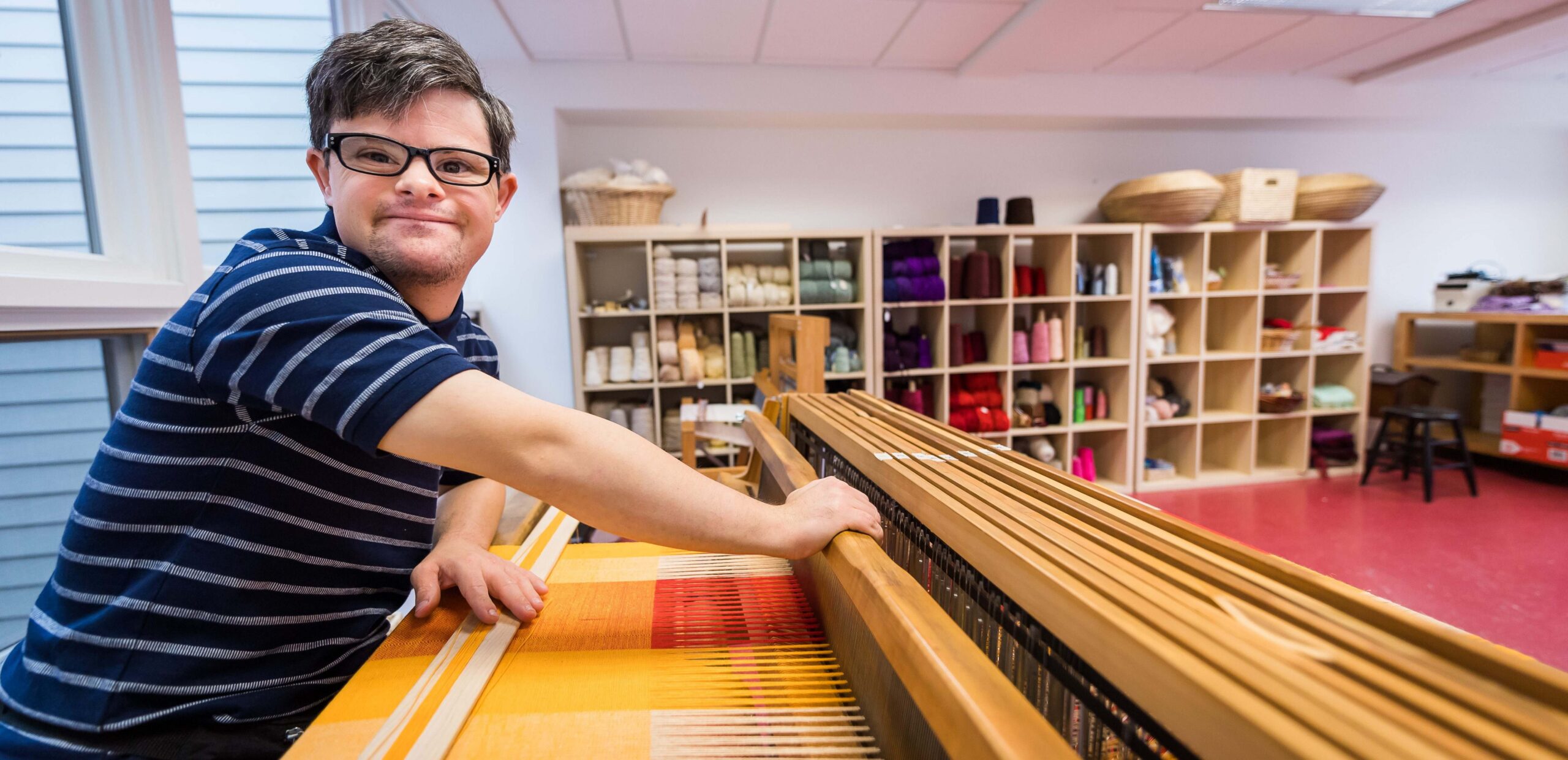 A young man weaving a bright yellow-and-orange pattern on a wide floor loom.