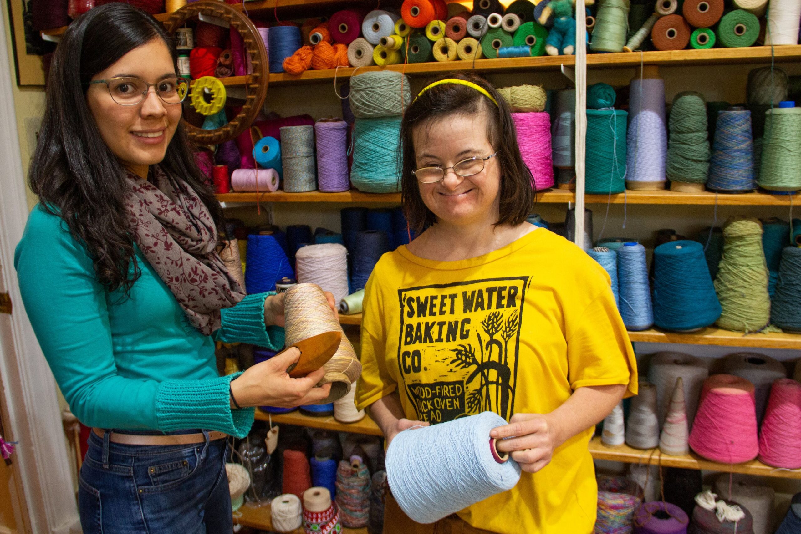 Two young women hold up cones of yarn, in front of sheles filled with more yarn and craft supplies.
