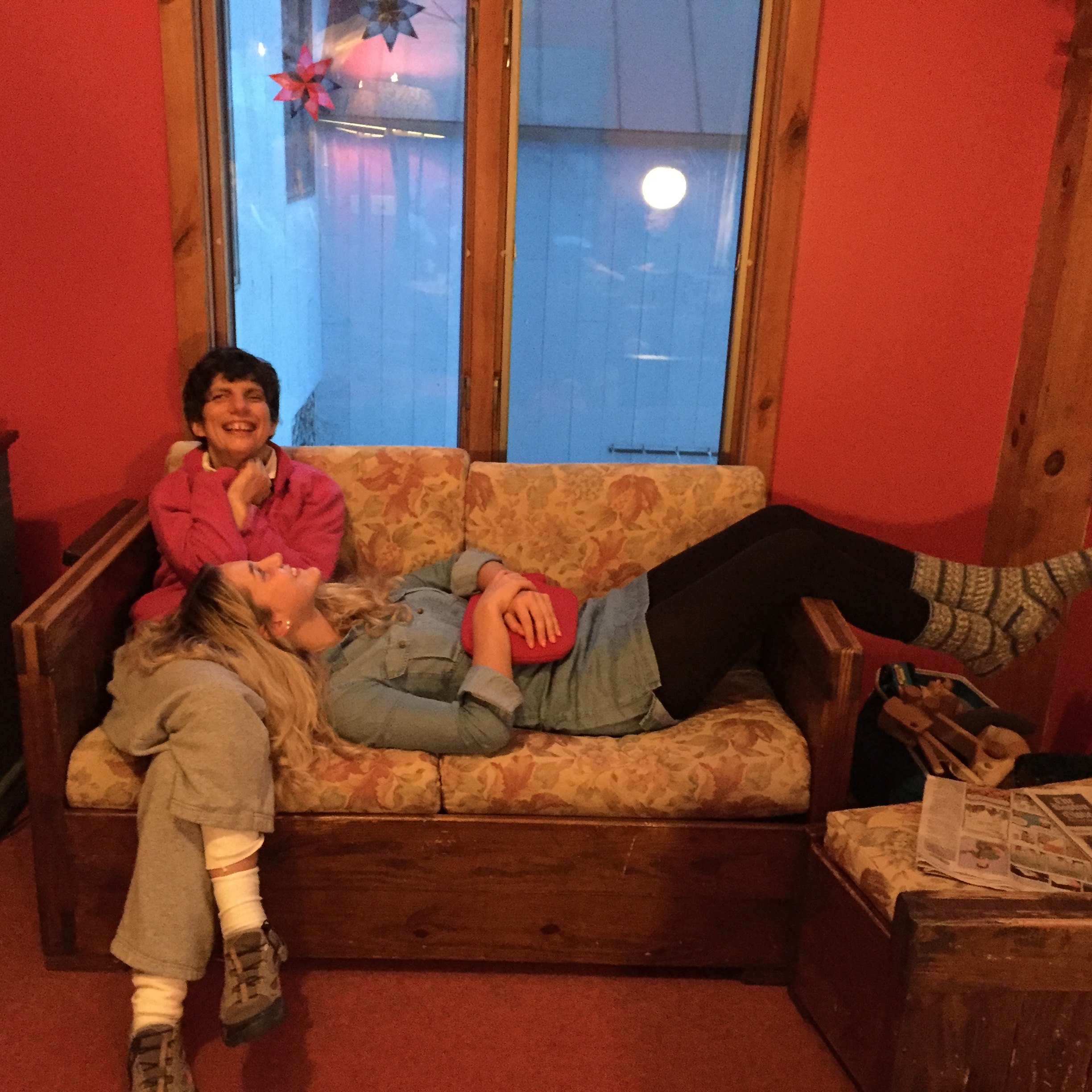 A young woman lies down on a couch with her head in an older woman's lap, both smiling and laughing.