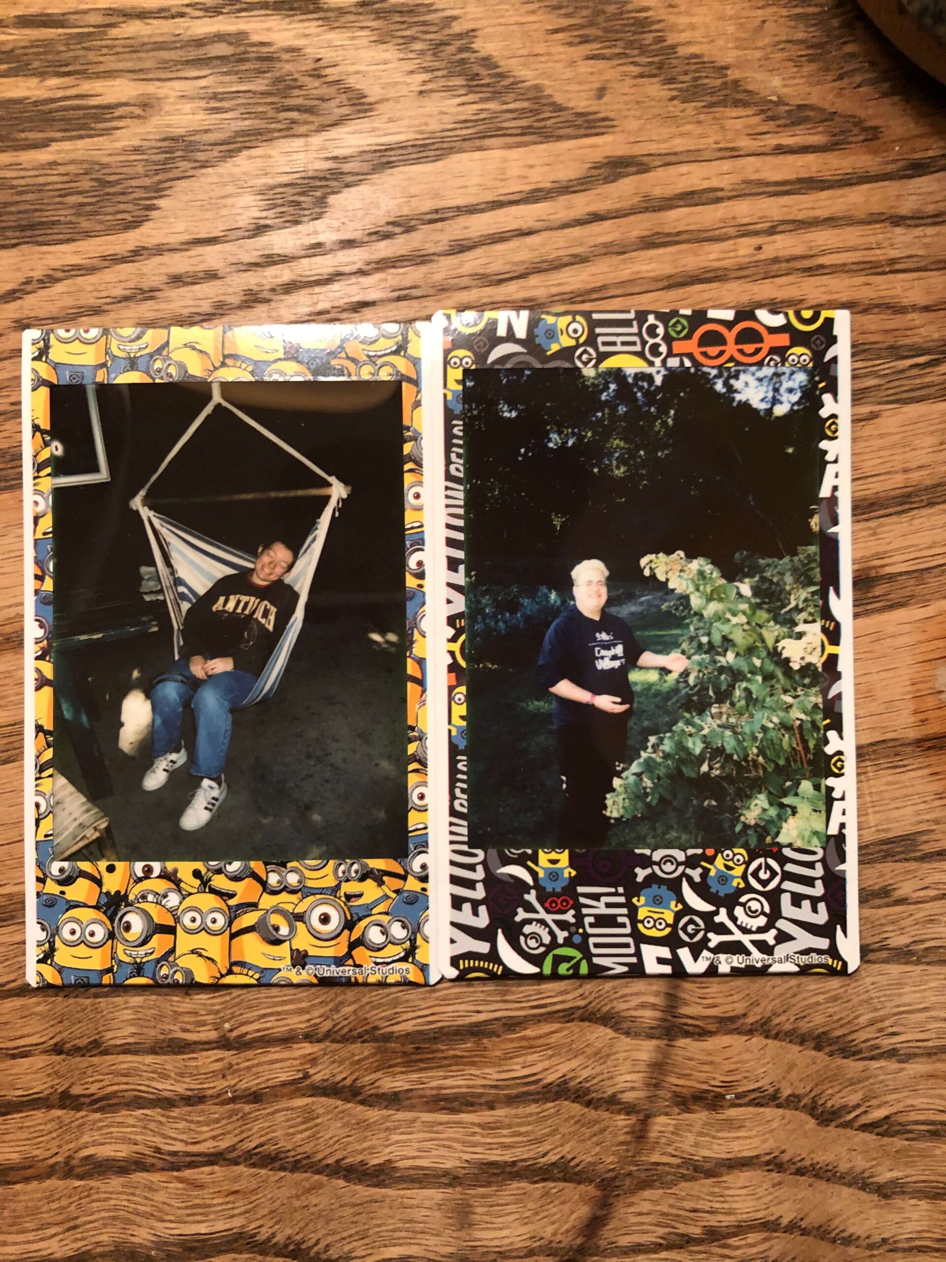 Two polaroids lying upon a wooden table. One is of a Delaney smiling in a hammock chair in the dark. The other is of a Asher with his hand inside of a bush with trees in the background. Both people are smiling at the camera. The polaroid pictures are decorated with Minion characters from the film Despicable Me.