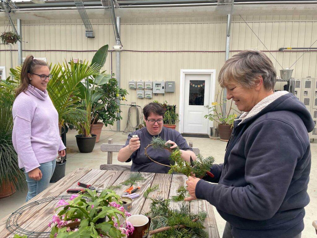 Three people are at a table inside of a greenhouse. The person in the middle is sitting and the two on the sides are standing. The person on the left is wearing lightwash jeans, a light purple turtleneck, black glasses, and their long brown hair in a high ponytail. The person in the middle is wearing a black tshirt, black glasses, and has short brown hair. The person on the right is wearing a black hooded jacket and has short grey hair. They are all smiling, and the person in the middle and right are working on creating wreaths, which they are looking at. Wreath-making supplies are laid out on the wooden table before them, including a wire frame, branches, flowers, and pruning shears.