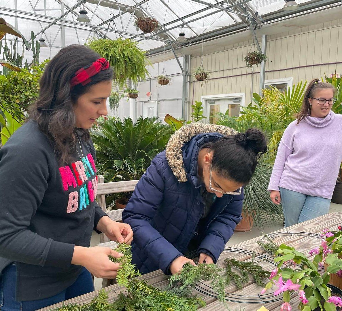 Three people are at a table inside of a greenhouse. the person on the left is wearing jeans, a grey sweater that says "merry and bright" in bright colors, and a red headband. They have long black hair. The person in the middle is wearing a blue puffy coat, clear glasses, and their long black hair is in a bun. The person on the right is wearing lightwash jeans, a lavender turtleneck sweater, black glasses, and their long brown hair in a high ponytail. Wreath-making supplies are laid out on the wooden table before them, including a wire frame, branches, flowers, and pruning shears. Behind them is a large collection of plants.