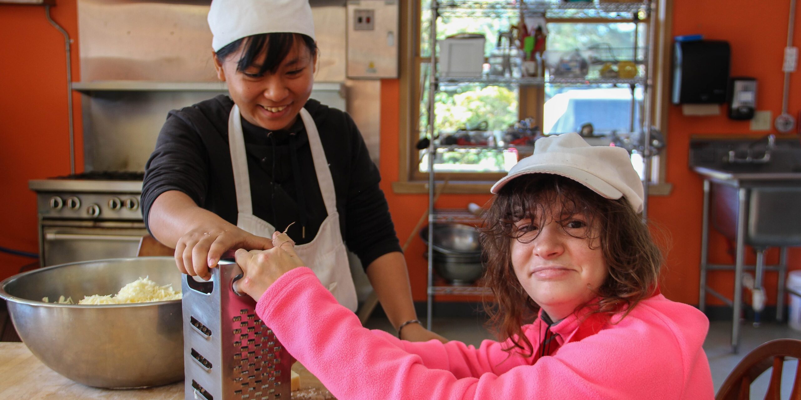 Two people working together in a cheerful red kitchen, grating cheese into a large bowl