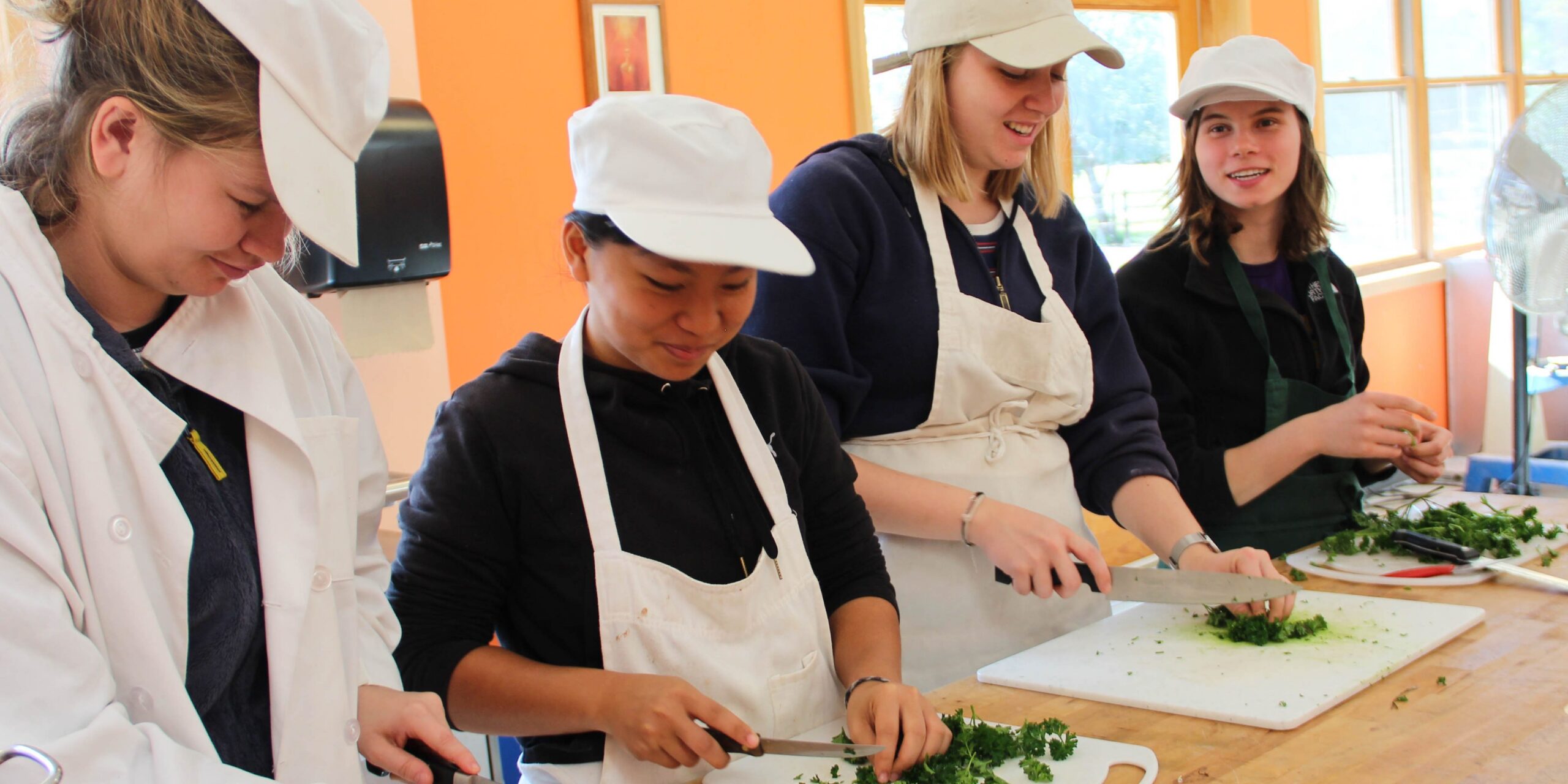 Four people in white hats and aprons chop up vegetables together on a wooden table.