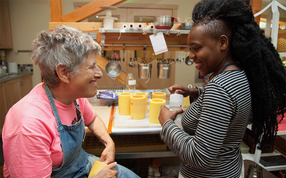 Two women smile at each other at a workbench where they are pouring candles.